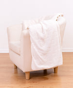 Pusheen Patterned Sherpa Blanket draped on top of a cream chair. The cream-colored blanket features a repeating outline of a graphic of Pusheen the Cat lounging. The large blanket is a soft polyester on one side while the reserve is a soft sherpa material. 