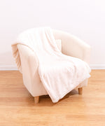 Cream-colored blanket is folded and draped over a cream-colored armchair. The large blanket has a repeating pattern of Pusheen the Cat embossed on the polyester material.  