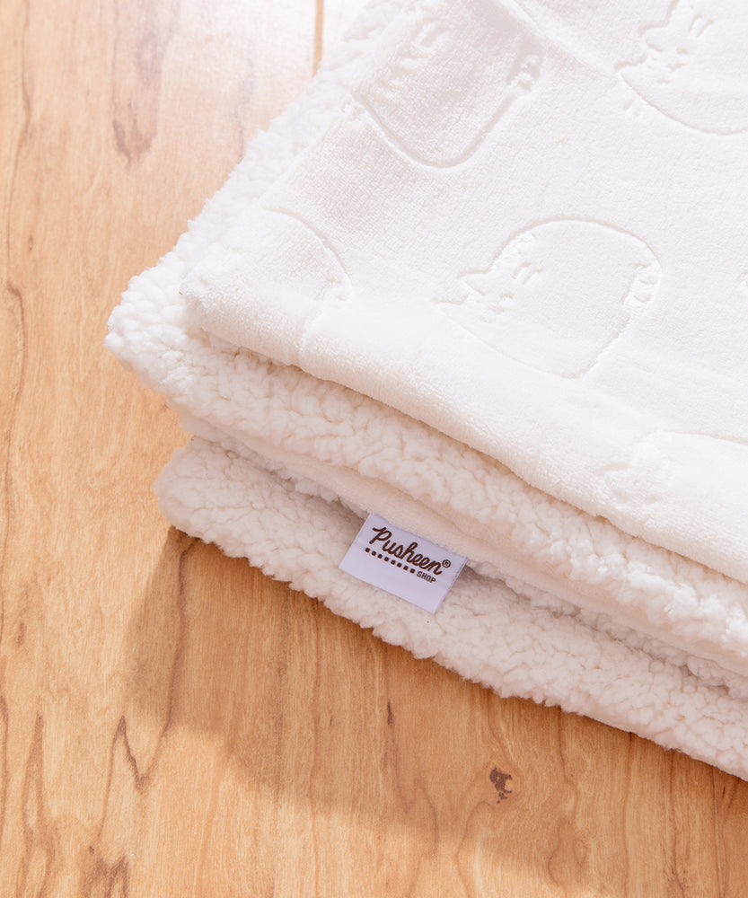 Close-up view of the Pusheen blanket. The white and brown Pusheen Shop logo tag is show on the edge of the polyester and sherpa blanket.  