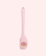 Close-up of included pink spatula in the set. At the scoop end of the spatula is a printed graphic of a pink glazed donut with multi-color sprinkles.