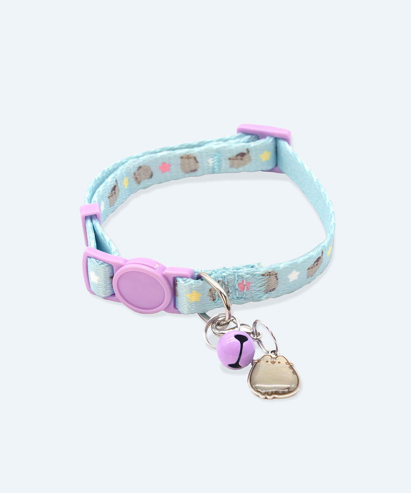 Close-up view of the Pusheen Pet Collar. The light blue collar has a print of multiple Pusheen poses and white, yellow, and pink stars in between the cats. The buckles and push-button opening mechanism are both light purple. Attached to the silver loop is a purple bell and Pusheen the Cat charm.