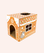 The other front quarter side view of the Pusheen Pet Gingerbread House Scratcher. This side features a graphic of Pusheen piping frosting on the gingerbread house. This side of the box has a hole that resembles a paned window.