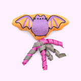 Close-up of the orange and purple bat-shaped plush pet toy with pink and grey ribbons attached to the bottom. 