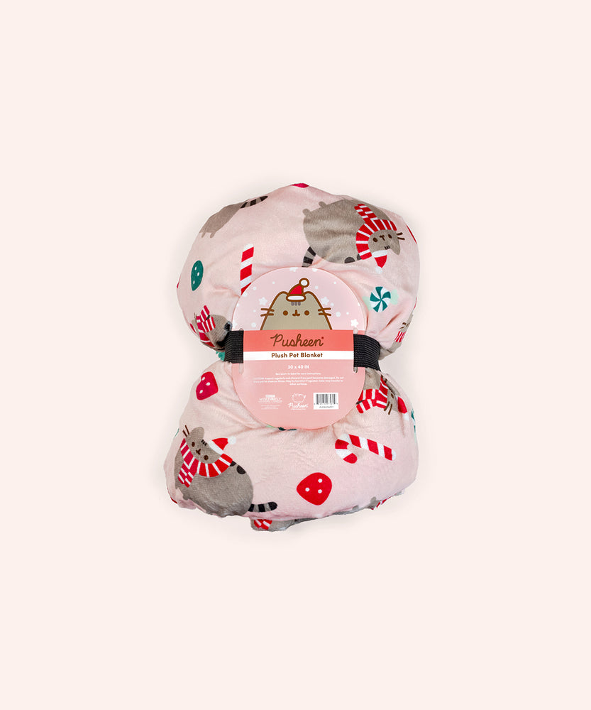 The Holiday Pet blanket is rolled inside its packaging which states that it is a “Pusheen Plush Pet Throw Blanket.” The packaged pink blanket is held together by a dark colored ribbon.