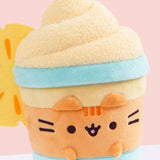 Close-up of Pineapple Float Pusheen. The medium-orange body is accompanied by blue horizontal lines to imitate a classic drink container. Pusheen's head stripes, eyes, smiling, mouth and whiskers are embroidered on the front of the plush while her orange ears and paws extend off the plush body.