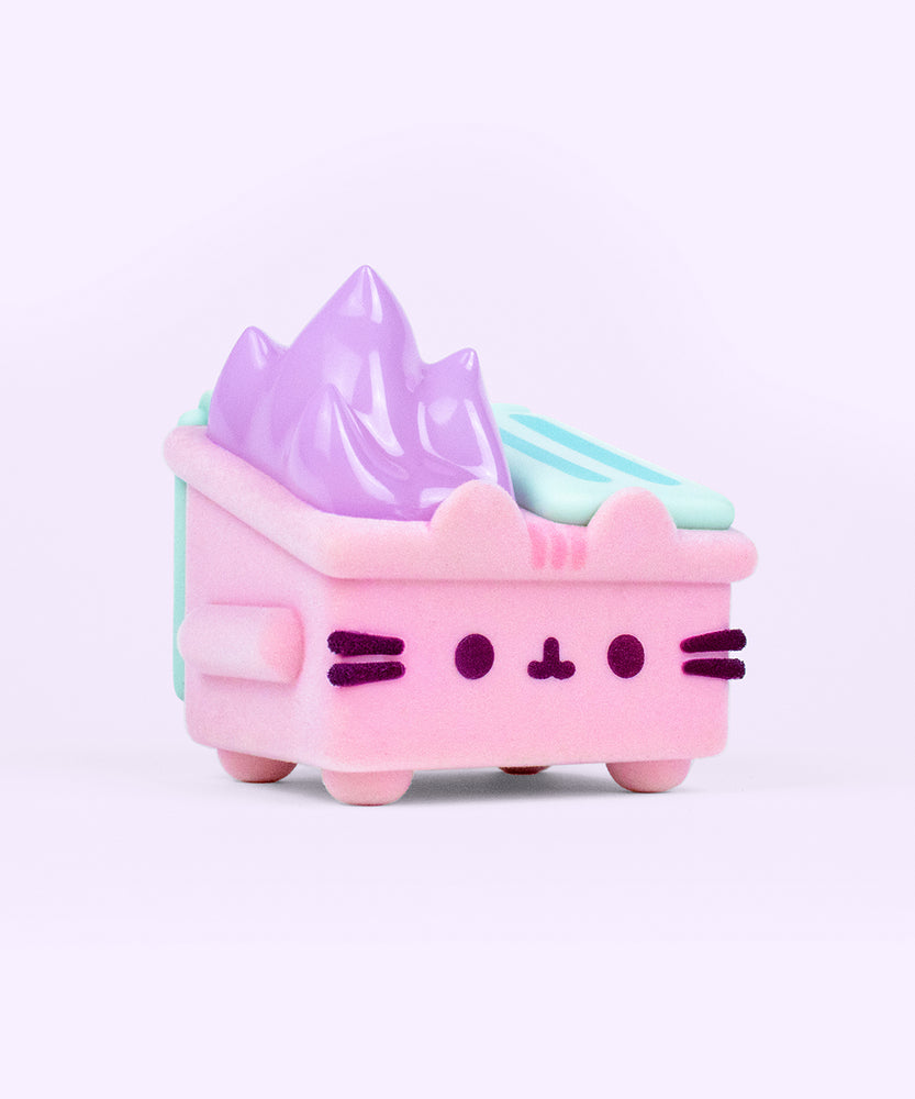 Front and side view of the Pusheen toy. The light pink fuzzy dumpster sits on four legs, has side handles, and mint green top lids. On the front of the toy is Pusheen’s face in dark purple fuzzy, 3D flocking. Coming out of the top of the toy figure is pastel purple flames. 