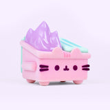 Front and side view of the Pusheen toy. The light pink fuzzy dumpster sits on four legs, has side handles, and mint green top lids. On the front of the toy is Pusheen’s face in dark purple fuzzy, 3D flocking. Coming out of the top of the toy figure is pastel purple flames. 