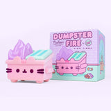Front view of Pusheen Pink Dumpster Fire Vinyl Figure. The fuzzy light pink figurine features Pusheen the Cat taking the form of a dumpster on fire. The pink body has Pusheen’s facial features in a dark purple while the mint green trash lid is accompanied by pastel purple flames. In the background is the figurine packaging featuring the figure design and pastel pink, mint green and purple details. 