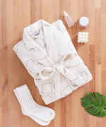 Cream bathrobe with cat design folded and surrounded by matching cream socks, a candle, and skincare products.