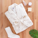 Cream bathrobe with cat design folded and surrounded by matching cream socks, a candle, and skincare products.