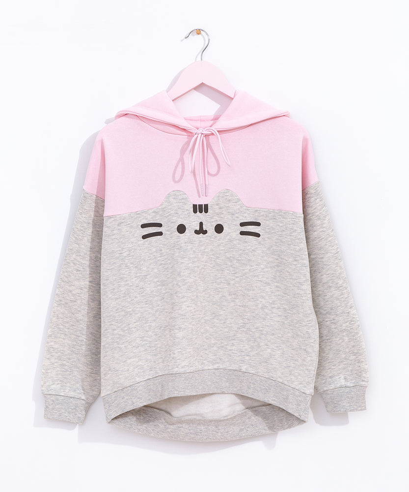 Pusheen Relaxed-Fit Character Hoodie Unisex hangs on a light pink hanger in front of a white wall. The two-toned sweatshirt features Pusheen’s face in the center of the hoodie. The top half of the hoodie is light pink while the bottom portion is light gray and outlines Pusheen’s rounded triangle ears to separate the two colors. 