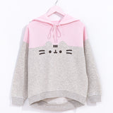 Pusheen Relaxed-Fit Character Hoodie Unisex hangs on a light pink hanger in front of a white wall. The two-toned sweatshirt features Pusheen’s face in the center of the hoodie. The top half of the hoodie is light pink while the bottom portion is light gray and outlines Pusheen’s rounded triangle ears to separate the two colors. 