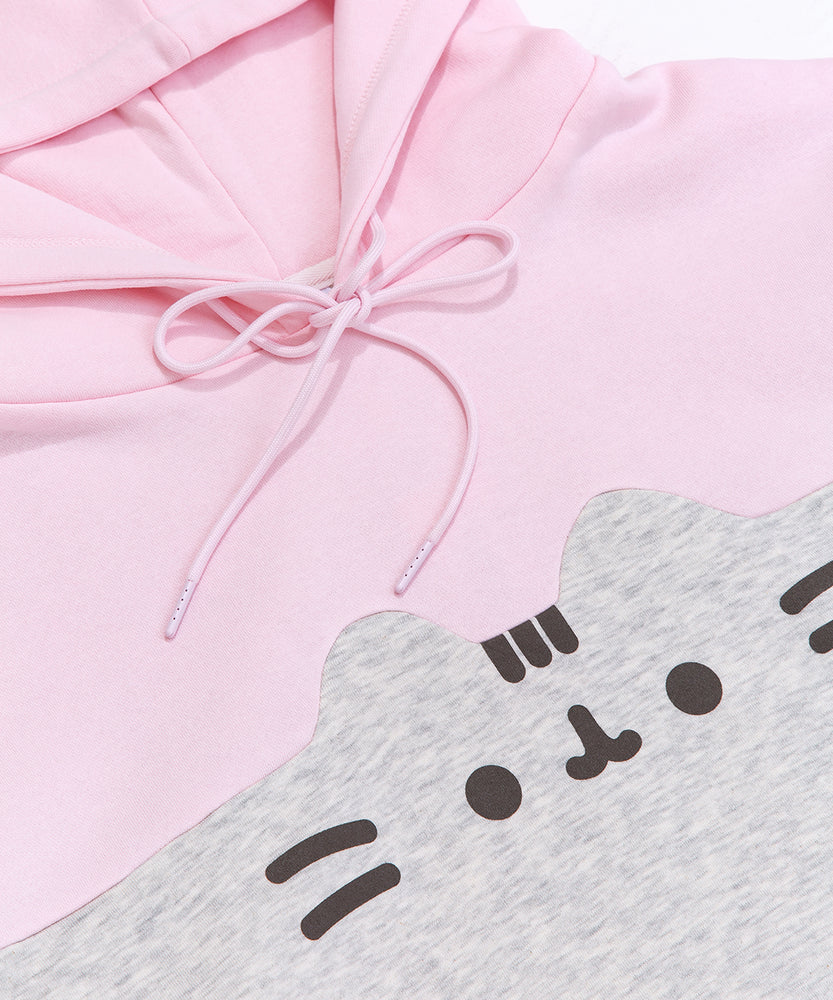 A close-up view of the printed graphic on the front center chest of the sweatshirt. The classic Pusheen pose shows her whiskers, mouth, eyes,head stripes in a dark gray print. The light pink hoodie drawstring is tied in a bow.  