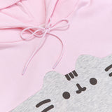 A close-up view of the printed graphic on the front center chest of the sweatshirt. The classic Pusheen pose shows her whiskers, mouth, eyes,head stripes in a dark gray print. The light pink hoodie drawstring is tied in a bow.  