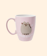 Front view of the Pusheen Self-Care Mug. The rounded light pink mug has a light pink handle with white interior walls. The outside of the mug features Pusheen the Cat grooming her paw surrounded by sparkles and the slogan “self-care.” 