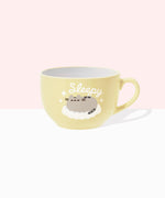 Front view of the Pusheen Sleepy Mug. The rounded light-yellow mug is perfect for lattes. The exterior has a light-yellow handle with white interior walls. The outside of the mug features Pusheen the Cat sleeping on a fluffy, white cloud surrounded by sparkles and the slogan “sleepy.” 