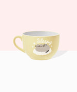 Back view of Pusheen latte mug. The light-yellow cup features a grey and brown graphic of Pusheen sleeping on a white, puffy cloud. Around Pusheen are white sparkles and above the cat is the phrase “sleepy” in white print. 