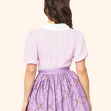 Back view of the purple blouse. The oversized white collar extends to the back of the blouse. The blouse has oversized tulip-like sleeves that end at the wearer's elbows.