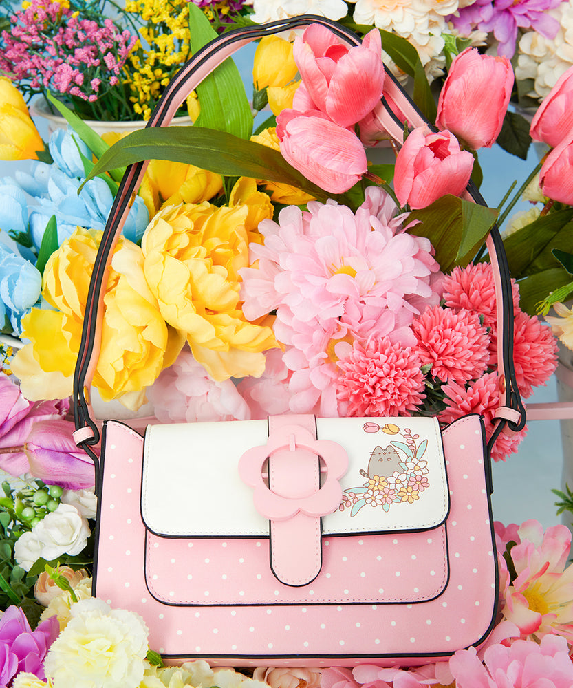 Pusheen Purse in a bed of fake flowers. The light pink purse has white polka dots, a pink daisy buckle, and a graphic of Pusheen surrounded by flowers on one corner of the closure flap.