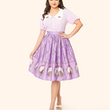 Front view of purple retro-inspired skirt featuring flowers and hearts in a diamond print and portraits of Pusheen the Cat, Little Sister Stormy, and Little Brother Pip at the bottom hem. The model’s hand holds skirt fabric away from the body to show off the skirt pattern.
