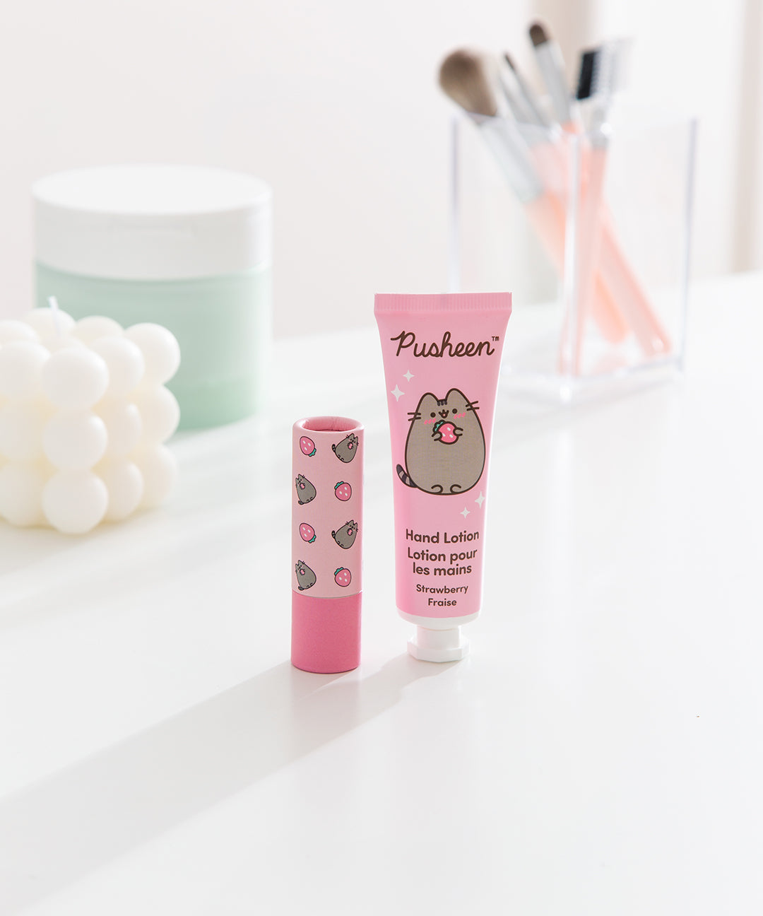Pusheen strawberry hand lotion and unscented lip balm sit on a white desktop. The set has pink packaging with strawberry graphics. 
