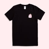 A black graphic short sleeve tee against a pink background. The graphic features Pusheen and a small strawberry milk carton printed on the wearer’s left chest.