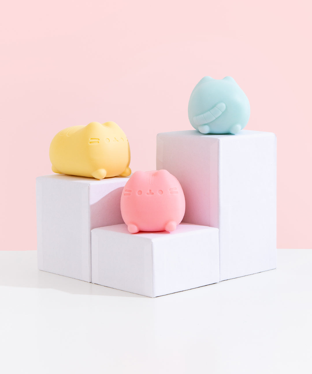 Pusheen Surprise Squishu Assortment including yellow, blue and pink colors on white pedestals. The front and side can be seen of the yellow squishy. The back of the blue squishy can be seen. Lastly, the front of the pink squishy toy can be seen.