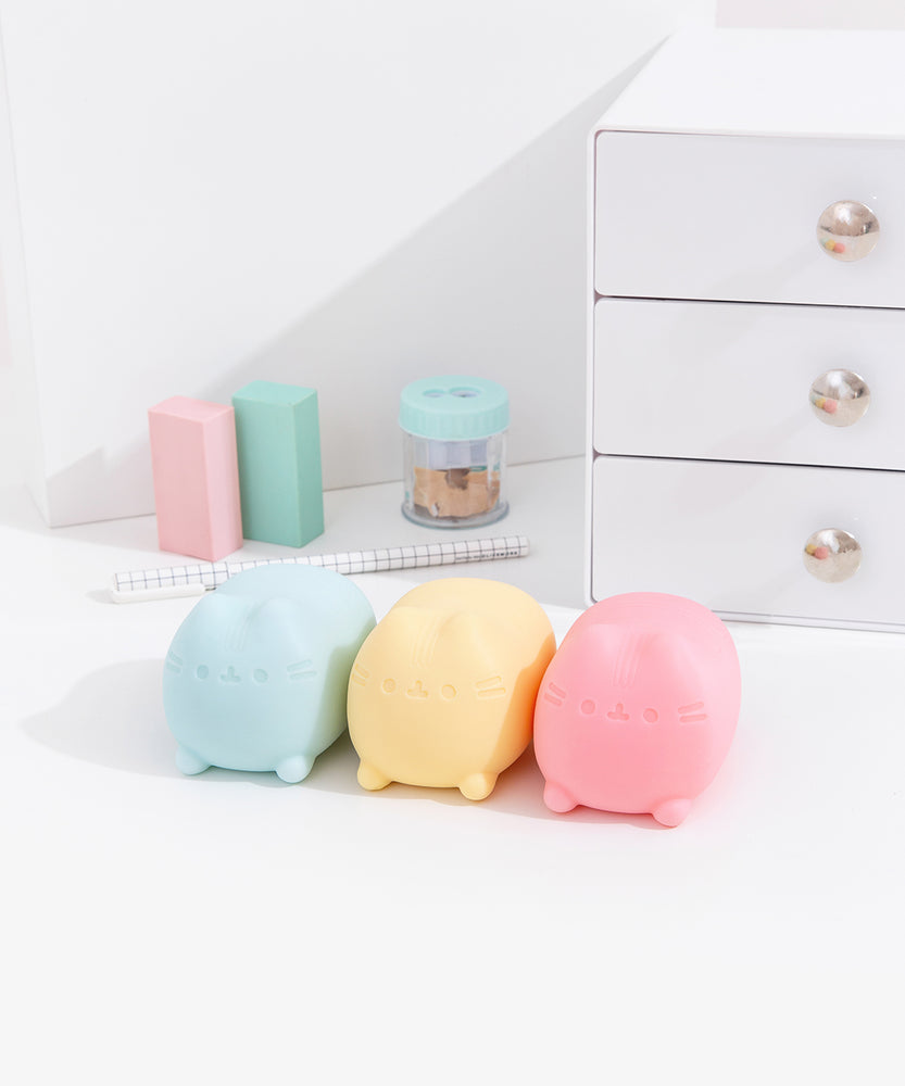 Blue, yellow, and pink Pusheen toys sit in front of stationery and desk items.