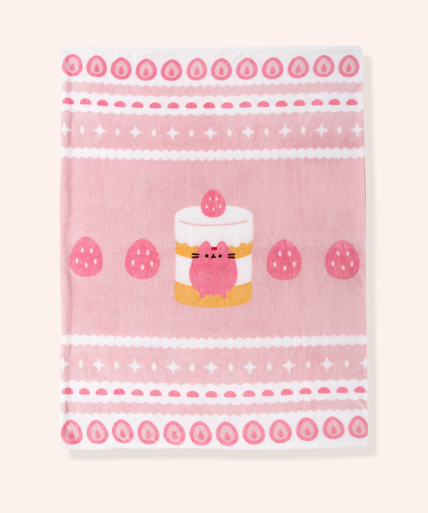 Full view of Pusheen Blanket. The polyester fabric features repeating graphics of strawberries, stars, and gum drops on the top and bottom of the blanket. The middle of the blanket has a graphic of a large layered cake with strawberries next to it.