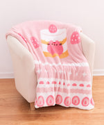 Pink-colored blanket is folded and draped over a cream-colored armchair. The large blanket has a repeating pattern of sweets graphics around the border with a large graphic of Pusheen the Cat in the very middle.