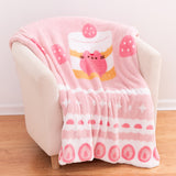 Pink-colored blanket is folded and draped over a cream-colored armchair. The large blanket has a repeating pattern of sweets graphics around the border with a large graphic of Pusheen the Cat in the very middle.