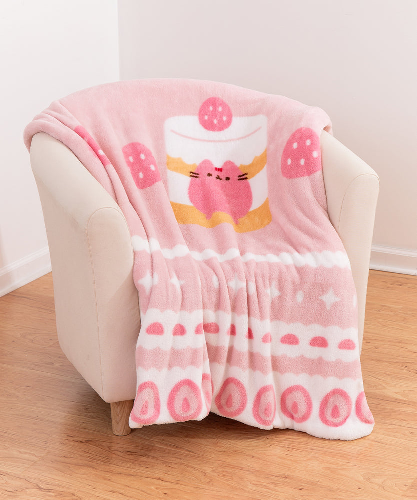 Pusheen Sweets blanket rests on a chair. The center graphic shows a pink Pusheen sitting inside a yellow and white layered cake with a pink strawberry on top.