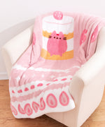 Pusheen Sweets Blanket draped on top of a cream chair. The pink blanket features a graphic of the Strawberry Sweets Cake Pusheen in the middle with a variety of sweets icons around the border. The large blanket is a soft polyester on both sides. 