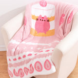 Pusheen Sweets Blanket draped on top of a cream chair. The pink blanket features a graphic of the Strawberry Sweets Cake Pusheen in the middle with a variety of sweets icons around the border. The large blanket is a soft polyester on both sides. 