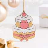 Pusheen Christmas Ornament hangs in front of gold holiday decorations. The strawberry layered cake ornament features Pusheen in pink inside the top layer of a yellow cake with white frosting.