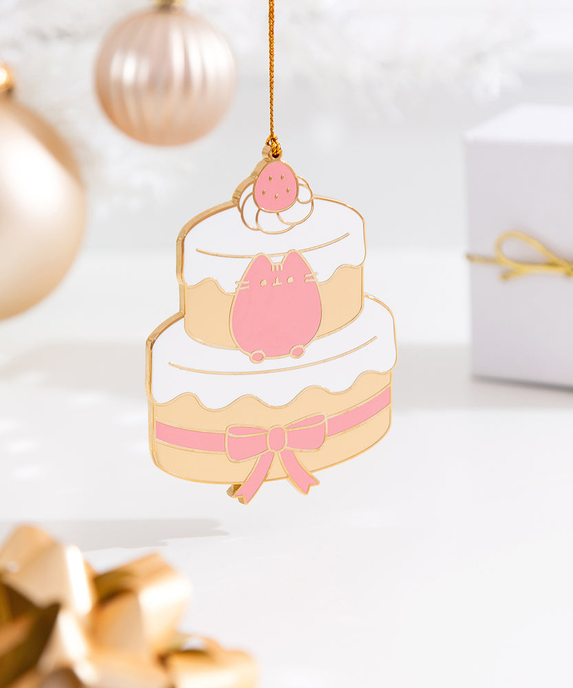 Strawberry cake ornament next to its white storage bag that features the Pusheen logo with the year the ornament launched. At the very top of the cake sits a whipped cream spot with a strawberry. The gold hanging string is attached to this strawberry topper. 