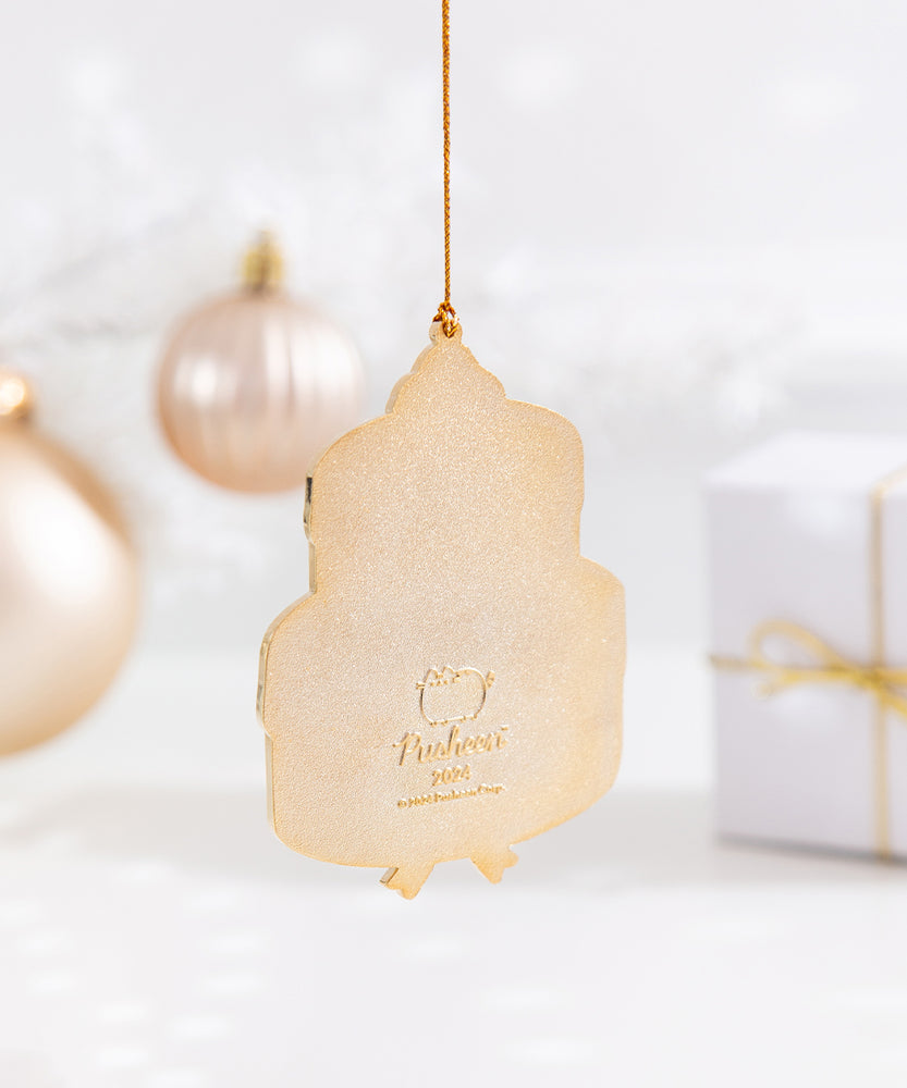 Back view of the ornament, which is a solid gold. The design on the back is the Pusheen the Cat logo and the copyright information. The cake shaped ornament hangs by its gold string in front of a Christmas ornaments and gift box. 