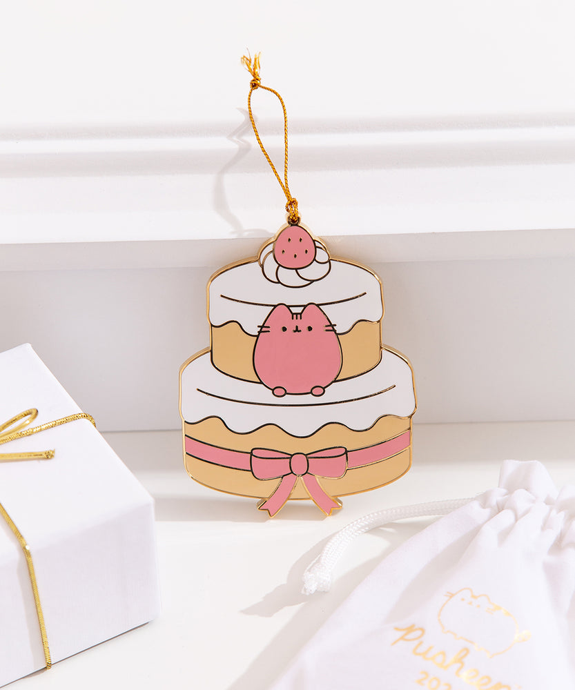 Pusheen Sweets Ornament rests on a white ledge. Pusheen is shown in pink and sits between the top and bottom layers of the strawberry cake.