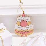 Pusheen Sweets Ornament rests on a white ledge. Pusheen is shown in pink and sits between the top and bottom layers of the strawberry cake.