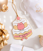 A flat ornament with a gold string lying next to a soft, white storage bag surrounded by a gold gift bow, yellow gold, light pink, and white ornaments. The ornament features Pusheen as as a layered dessert cake. The front of the ornament is in color and has gold outlines. 