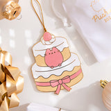 A flat ornament with a gold string lying next to a soft, white storage bag surrounded by a gold gift bow, yellow gold, light pink, and white ornaments. The ornament features Pusheen as as a layered dessert cake. The front of the ornament is in color and has gold outlines. 