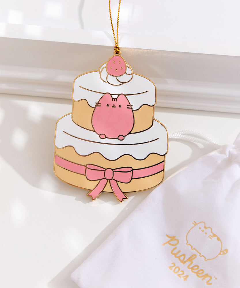 Strawberry cake ornament next to its white storage bag that features the Pusheen logo with the year the ornament launched. At the very top of the cake sits a whipped cream spot with a strawberry. The gold hanging string is attached to this strawberry topper. 