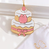 Strawberry cake ornament next to its white storage bag that features the Pusheen logo with the year the ornament launched. At the very top of the cake sits a whipped cream spot with a strawberry. The gold hanging string is attached to this strawberry topper. 