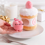 Model holds the removable Pusheen the Cat pink plush that sits inside the strawberry cake plush environment. The light plush rests on the model’s fingers.  