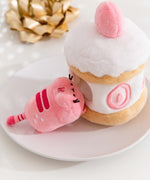 Side view of the Strawberry Plush. The removable interior plush rests in front of the cake environment. The little pink plush has white strawberry seeds embroidered on the booty area of the cat.  