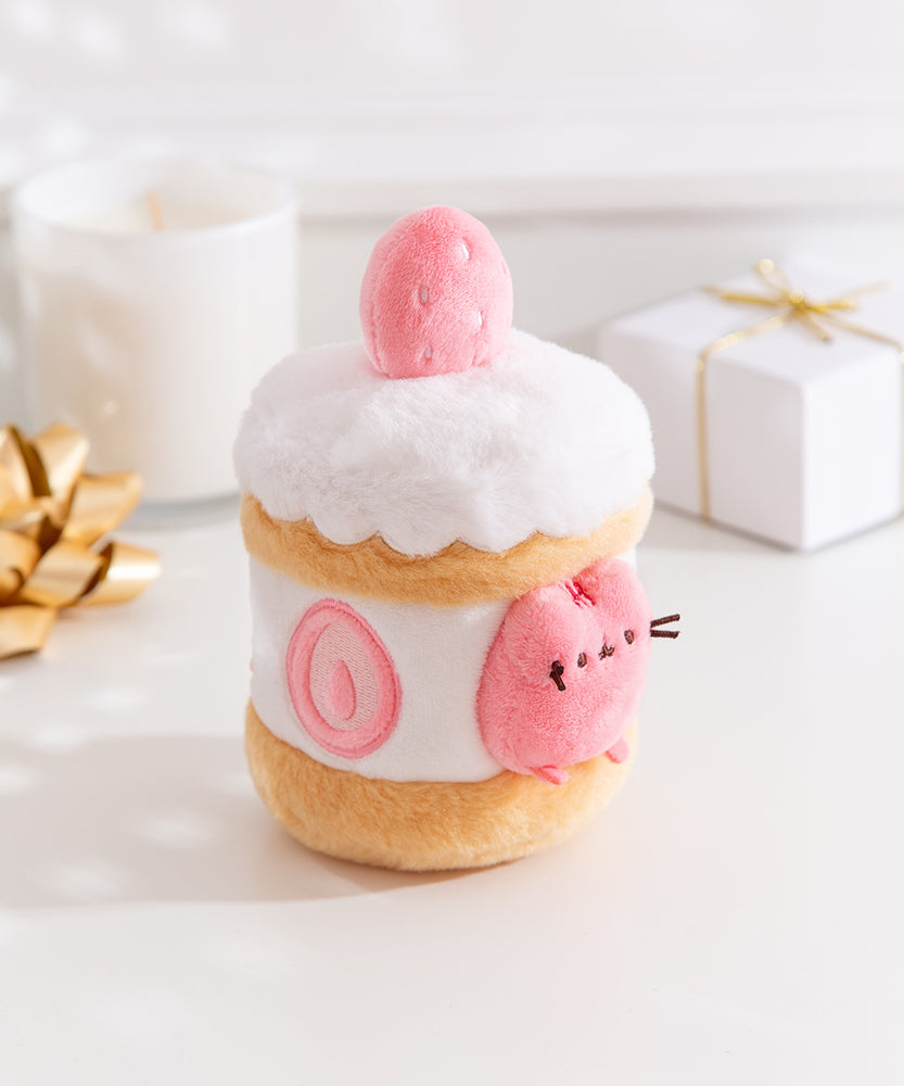 Side quarter view of the strawberry Pusheen plush. The cake plush has a light brown cake layer in the middle and at the bottom. The interior is a white icing layer with cut strawberries encircling the plush.  