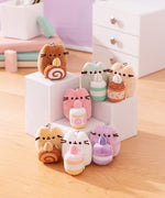 Front view of Pusheen Sweets Surprise Plush assortment. Plush keychains sit on multi-level white pedestal in front of a office desk set-up. Keychains feature Pusheen holding various sweet treats including a lavender macaron, swiss roll, caramel roll, and more.