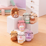 Front view of Pusheen Sweets Surprise Plush assortment. Plush keychains sit on multi-level white pedestal in front of a office desk set-up. Keychains feature Pusheen holding various sweet treats including a lavender macaron, swiss roll, caramel roll, and more.