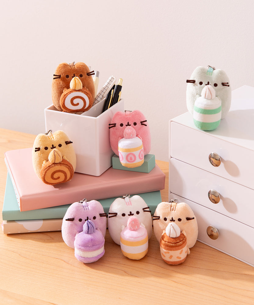 Alternate view of Pusheen Sweets Surprise Plush assortment. Plush keychains sit on multi-level pedestal in front of a office desk set-up. The mini plush Pusheens have a lobster claw clip for easy attachment to bags and zippers.