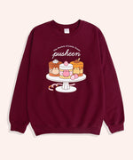 On a light backdrop lies a long sleeve burgundy graphic sweatshirt. The Pusheen Sweets Sweatshirt features a large graphic of Pusheen the Cat as three, layered cakes. The brown mocha cake on the left features chocolate sprinkles and is topped with whipped cream. The middle strawberry cake is layered with yellow cake and features cut strawberries throughout. The right caramel cake features caramel drinks off the top.  